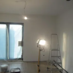 Wnętrze po malowaniu: białe ściany, zabezpieczone okno folią, drabina, lampa halogenowa i akcesoria malarskie na podłodze. Widoczny fragment ściany przed malowaniem.