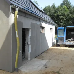 Budynek w trakcie ocieplania, z żółtą rurą wentylacyjną i dwoma niebieskimi samochodami dostawczymi w tle, ukazujący etap prac elewacyjnych.