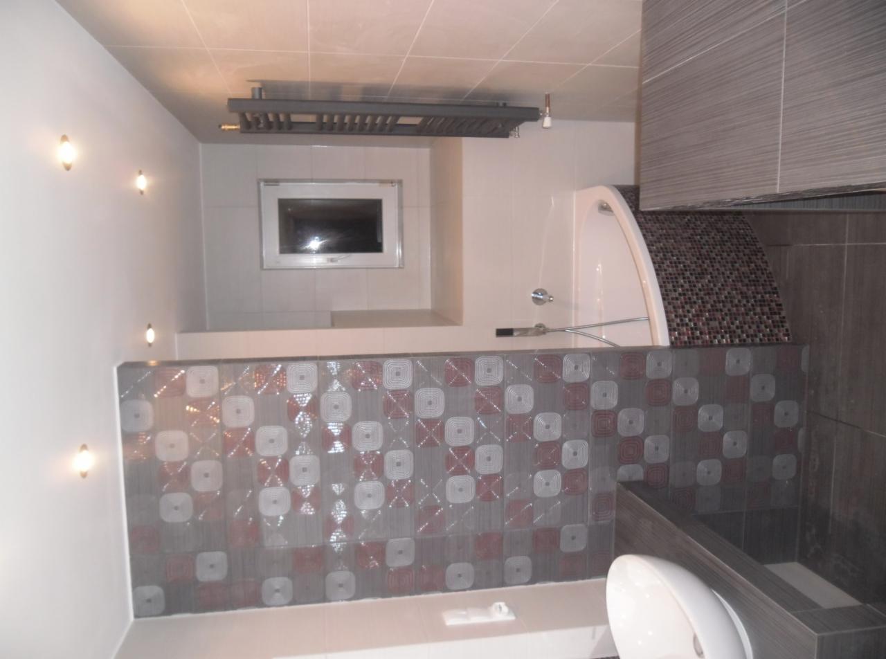 Wykończona łazienka z mozaiką na obudowie wanny, grzejnikiem drabinkowym i oświetleniem punktowym. Widok na umywalkę i toaletę w odcieniach szarości i bieli.