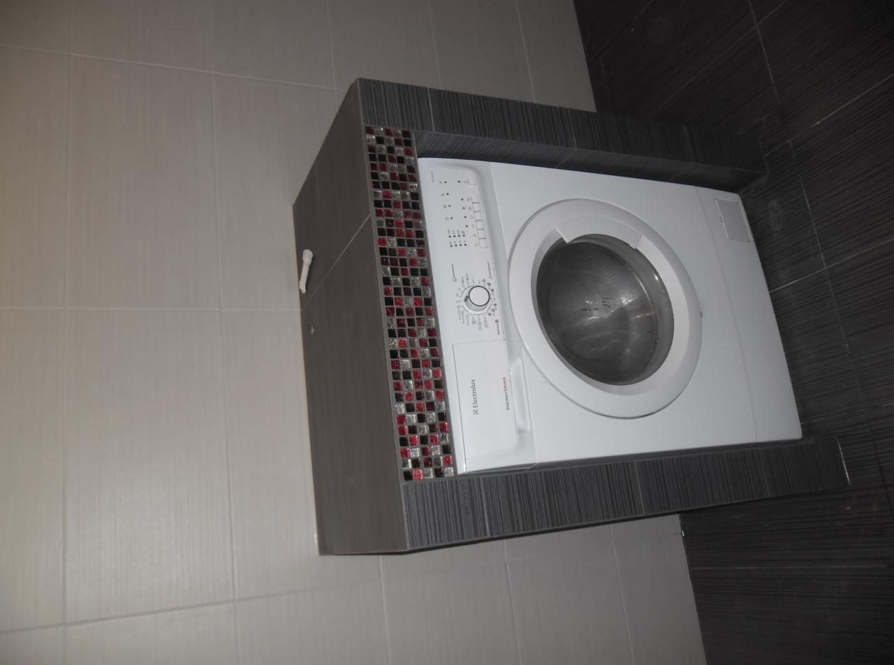 Biała pralka Electrolux zabudowana w szarej obudowie z mozaiką w czerwono-szarej kolorystyce, na tle płytek ściennych w łazience.