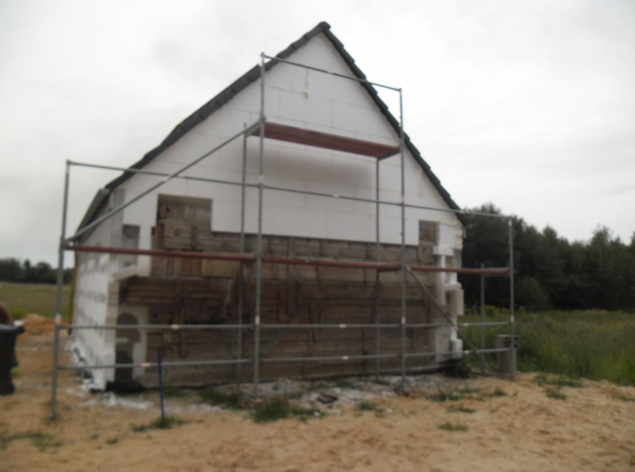 Dom w trakcie ocieplania: fragment elewacji z ociepleniem styropianowym, rusztowanie, widoczne fragmenty starej drewnianej konstrukcji. Tło: łąka, drzewa, pochmurne niebo.