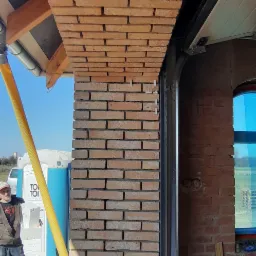 Fragment ściany z cegły klinkierowej w trakcie budowy garażu, widoczny pracownik i przenośna toaleta w tle.