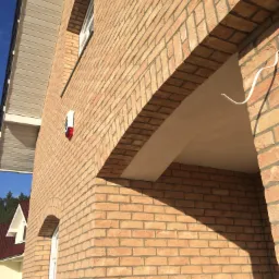 Ściana domu z jasnej cegły klinkierowej z łukowym wejściem, widoczny alarm na elewacji i fragment dachu sąsiedniego budynku.