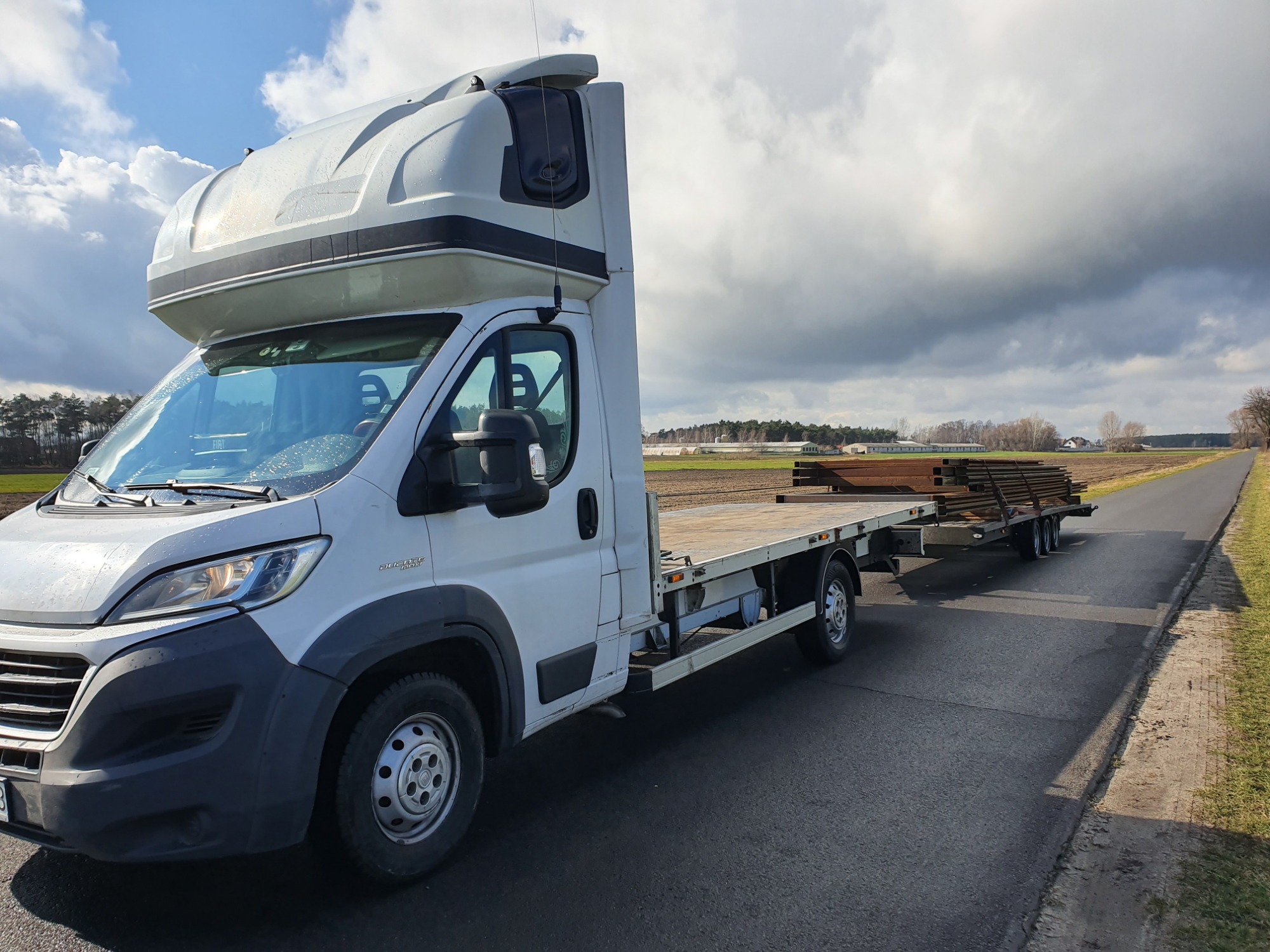 Biały samochód dostawczy z platformą i przyczepą przewożący drewniane elementy na asfalowej drodze, pochmurne niebo w tle.