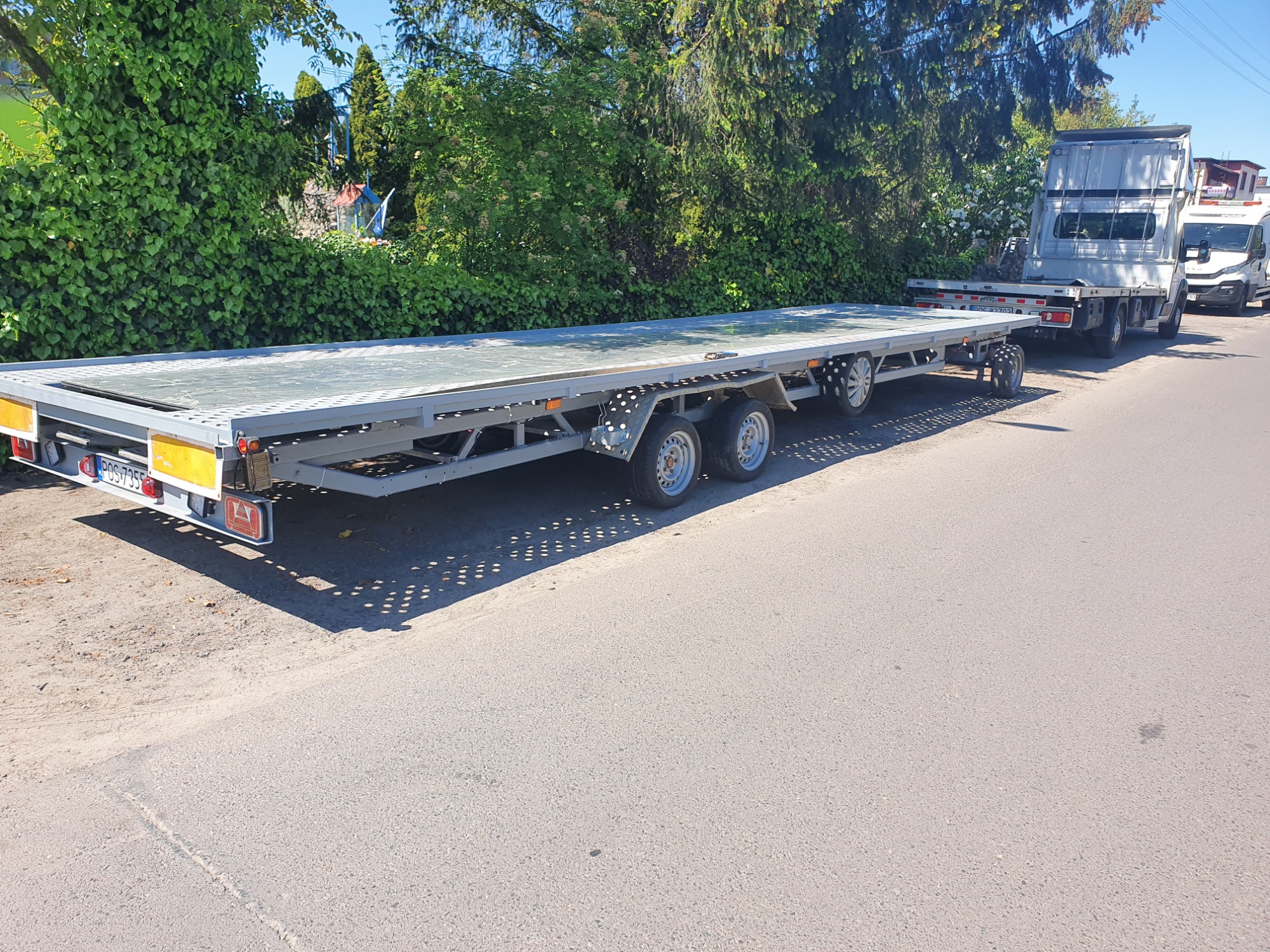 Długa, pusta laweta samochodowa z trzema osiami, zaparkowana na poboczu drogi w słoneczny dzień, podłączona do srebrnego samochodu ciężarowego.