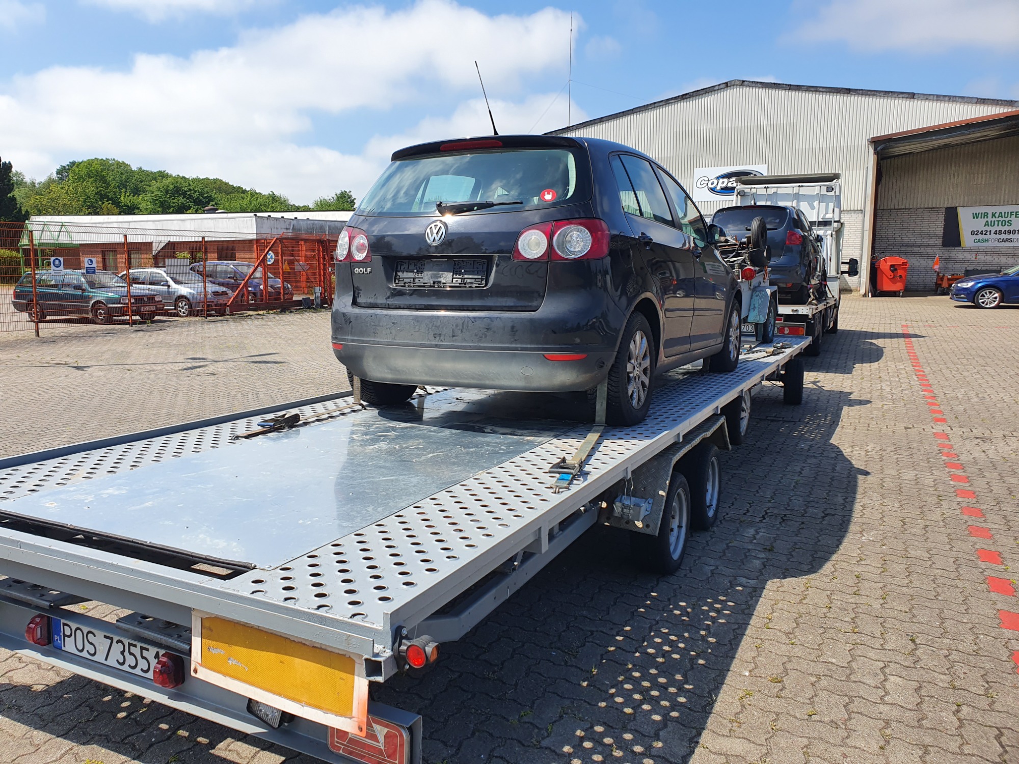 Czarny samochód VW Golf na srebrnej, perforowanej lawecie z polską tablicą rejestracyjną POS 7355, zaparkowanej na placu przed budynkiem firmy. Na lawecie za Golfem widoczny drugi, ciemny samochód.