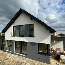 Dwukolorowa elewacja domu jednorodzinnego z dużymi oknami, widoczne materiały budowlane na placu budowy, pochmurne niebo.