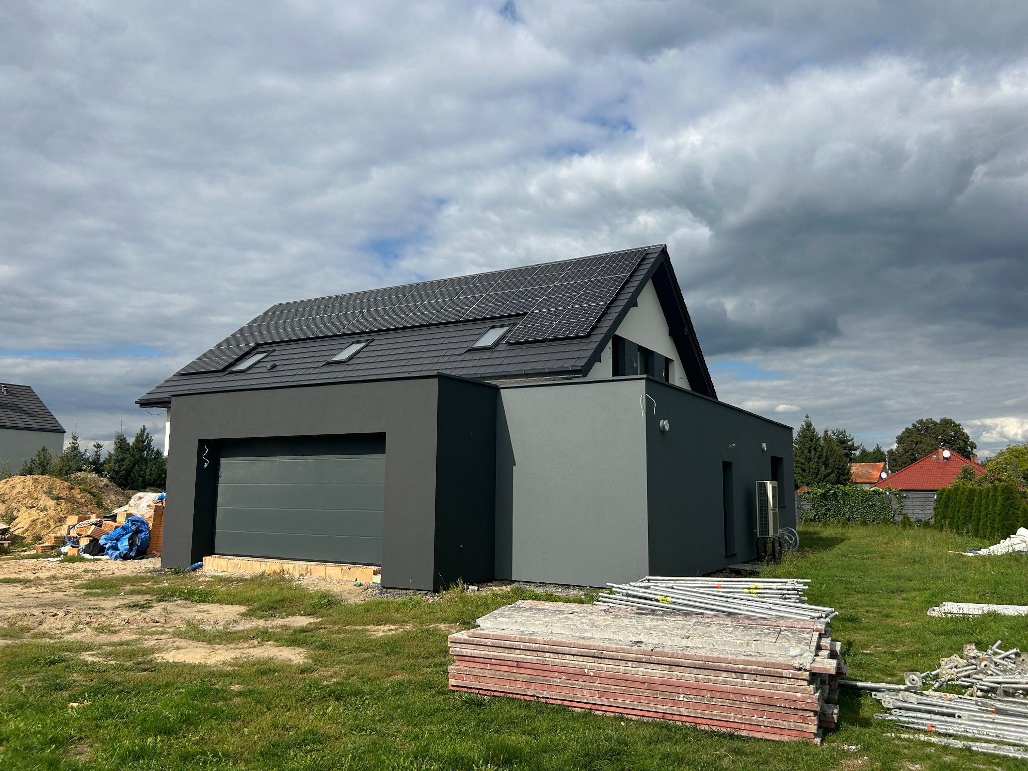 Nowoczesny dom z szarą elewacją i panelami słonecznymi na dachu, w tle pochmurne niebo, na trawniku leżą materiały budowlane i rusztowania.