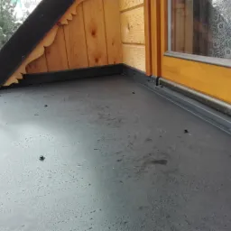 Nowe, czarne pokrycie dachu płaskiego z widocznymi śrubami mocującymi, drewnianą konstrukcją i oknem z koronkową zasłoną w tle.