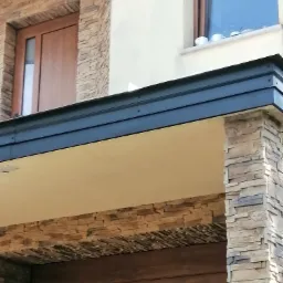 Fragment budynku z kamienną elewacją, ciemną blachą okapową i drewnianą stolarką okienną, z widocznymi filiżankami na parapecie okna.