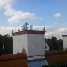 Biały komin z metalowym wykończeniem na dachu z czerwonej dachówki, na tle błękitnego nieba z chmurami. Widoczny wiatraczek na kominie.