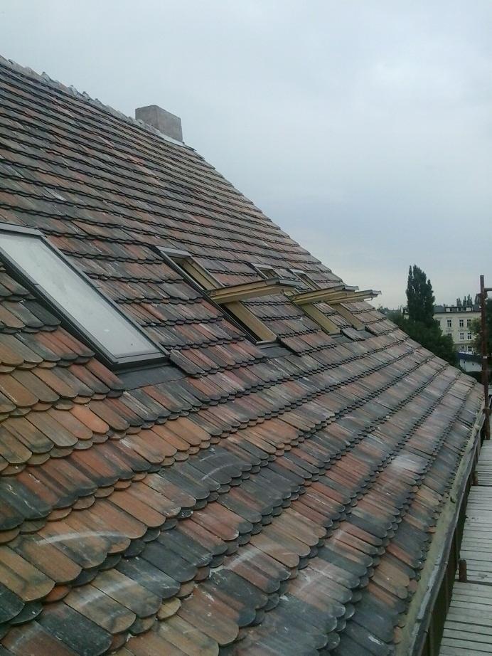 Dach z dachówki karpiówki w odcieniach brązu i czerni, z trzema oknami dachowymi i kominem w tle, widziany z boku pod kątem. Widoczne rusztowanie.