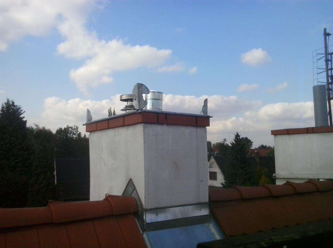 Biały komin z metalowym wykończeniem na dachu z czerwonej dachówki, na tle błękitnego nieba z chmurami. Widoczny wiatraczek na kominie.