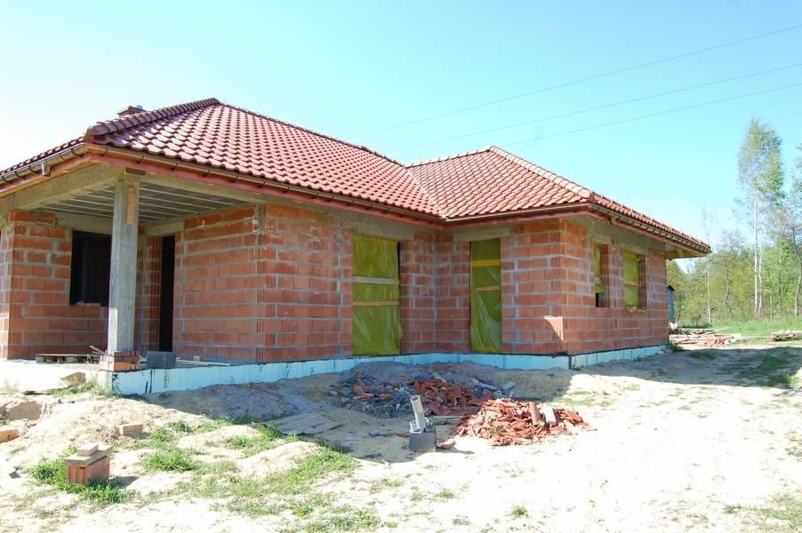 Dom w trakcie budowy: czerwona dachówka, murowane ściany z cegły, okna zabezpieczone folią. Widoczne materiały budowlane i otoczenie w słoneczny dzień.