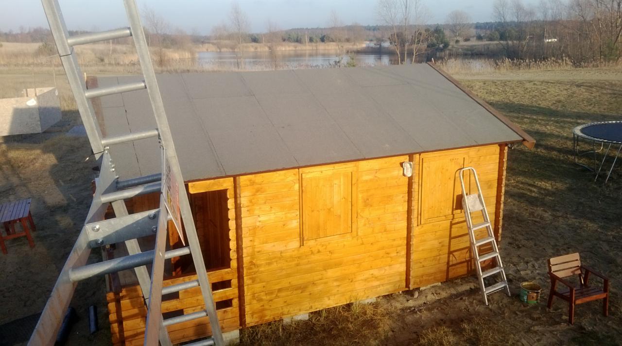 Drewniany domek z pokrytym papą dachem w trakcie budowy, z drabiny i otoczenia widać krajobraz jeziora. Druga drabina oparta o ścianę, obok krzesło i puszka.