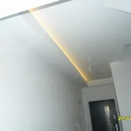 Minimalistyczne wnętrze: gładkie, białe ściany, sufit z oświetleniem LED wzdłuż krawędzi i wisząca żarówka. Widoczny fragment drzwi wejściowych.