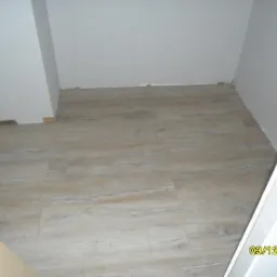 Jasne panele podłogowe imitujące drewno, ułożone w małym pomieszczeniu z białymi ścianami. Widoczny fragment drzwi i data wykonania zdjęcia.