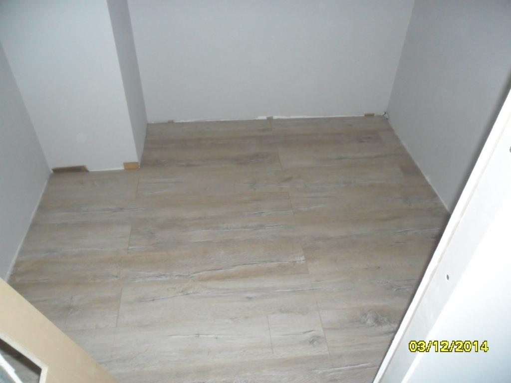 Jasne panele podłogowe imitujące drewno, ułożone w małym pomieszczeniu z białymi ścianami. Widoczny fragment drzwi i data wykonania zdjęcia.