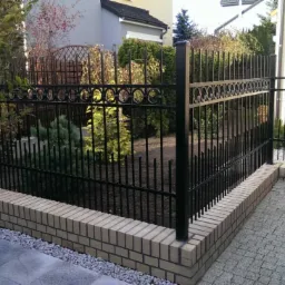 Czarne metalowe ogrodzenie z dekoracyjnymi okręgami, osadzone na niskim murku z jasnej cegły, przylegające do kostki brukowej i zaparkowanego czarnego samochodu.