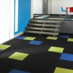 Aranżacja biura z ciemną wykładziną dywanową w kwadraty w odcieniach niebieskiego i limonki, schody metalowe prowadzące na podest z wykładziną, w tle kolorowe krzesła biurowe.