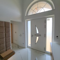 DREW-MEX Jarosław Bukowski - Nowoczesne wejście do domu z białymi drzwiami antywłamaniowymi z dekoracyjnym przeszkleniem, marmurową podłogą z ciemnymi żyłami i tapicerowanym panelem ściennym w odcieniach beżu.