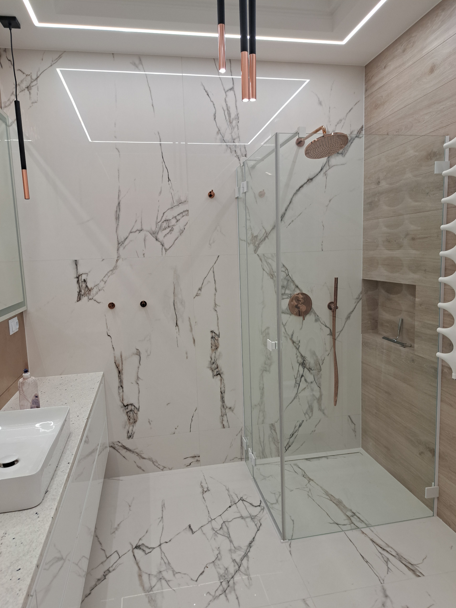 Nowoczesna łazienka z kabiną prysznicową typu walk-in, wykończona białymi płytkami imitującymi marmur z szarymi żyłami, armaturą w kolorze różowego złota i oświetleniem LED.