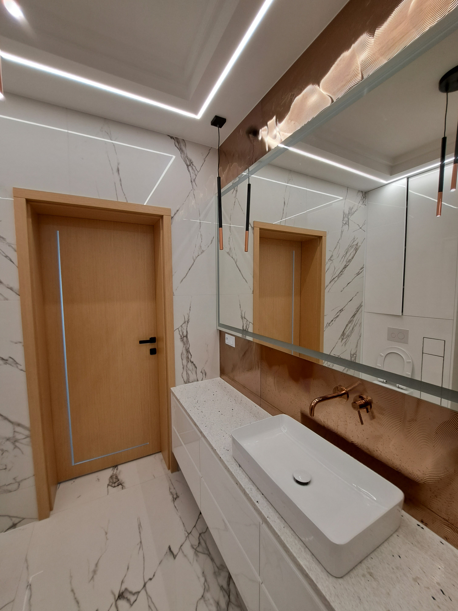 Nowoczesna łazienka z marmurowymi płytkami, drewnianymi drzwiami, białą umywalką nablatową, złotą armaturą i oświetleniem LED.