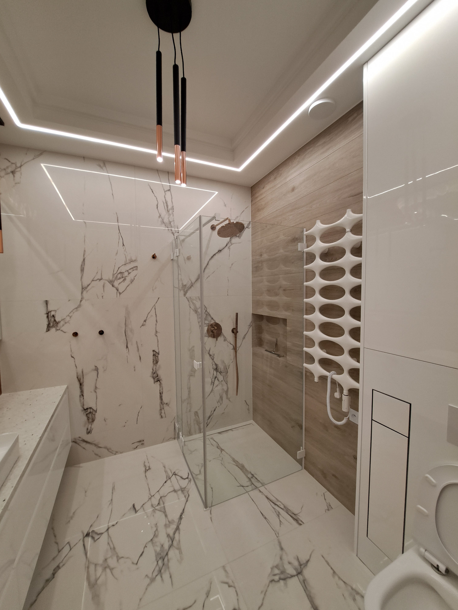 Nowoczesna łazienka z marmurowymi płytkami, szklaną kabiną prysznicową, grzejnikiem dekoracyjnym i podwieszaną toaletą, oświetlona listwami LED i wiszącymi lampami w kolorze czarnym i miedzianym.