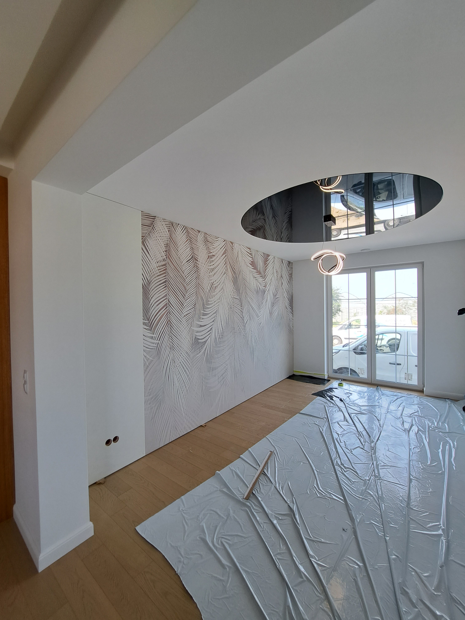 Pokój w trakcie prac, podłoga zabezpieczona folią, widoczna ściana z tapetą w liście, okrągły sufit podwieszany z lustrzanym wykończeniem i nowoczesną lampą.