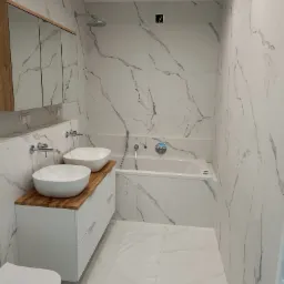 Nowoczesna łazienka z białymi płytkami imitującymi marmur, dwie umywalki nablatowe na drewnianym blacie, biała szafka z frezowanymi frontami, wanna w zabudowie, chromowana armatura i toaleta.