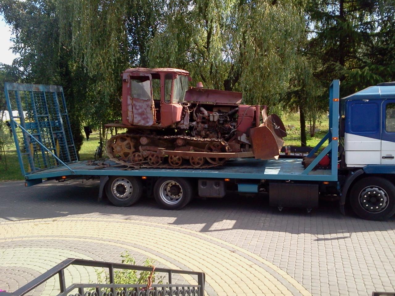 Czerwony, zardzewiały traktor gąsienicowy przewożony na niebieskiej platformie ciężarówki na tle drzew i kostki brukowej.