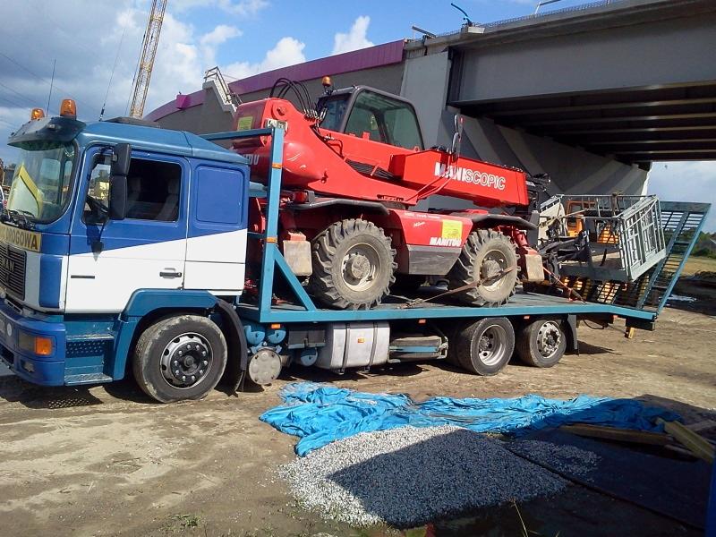 Laweta przewozi czerwoną ładowarkę teleskopową Manitou na tle konstrukcji mostu i placu budowy.