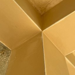 Mirosław Pagacz TYNK-SYSTEM - Świeżo nałożony, beżowy tynk maszynowy w rogu pomieszczenia, widoczne niedoskonałości na suficie i łączeniach ścian.