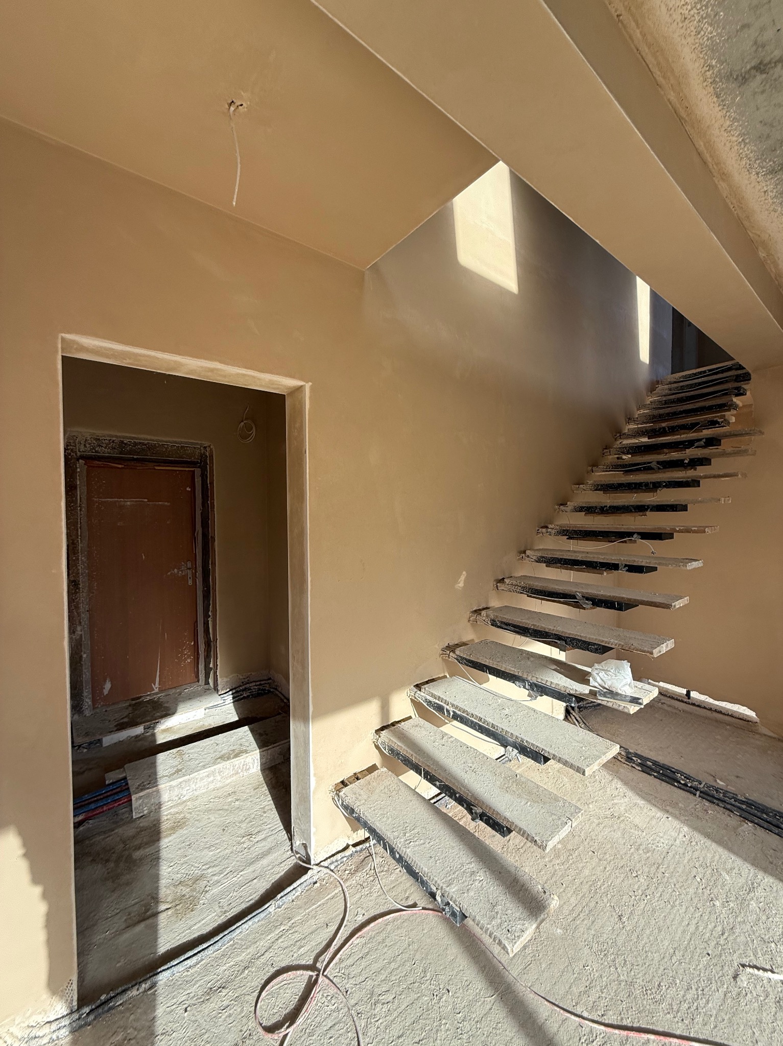 Wnętrze w trakcie prac wykończeniowych: świeżo otynkowane ściany w odcieniach beżu, surowe schody z widoczną konstrukcją metalową i betonowymi stopniami w trakcie montażu, kable elektryczne...