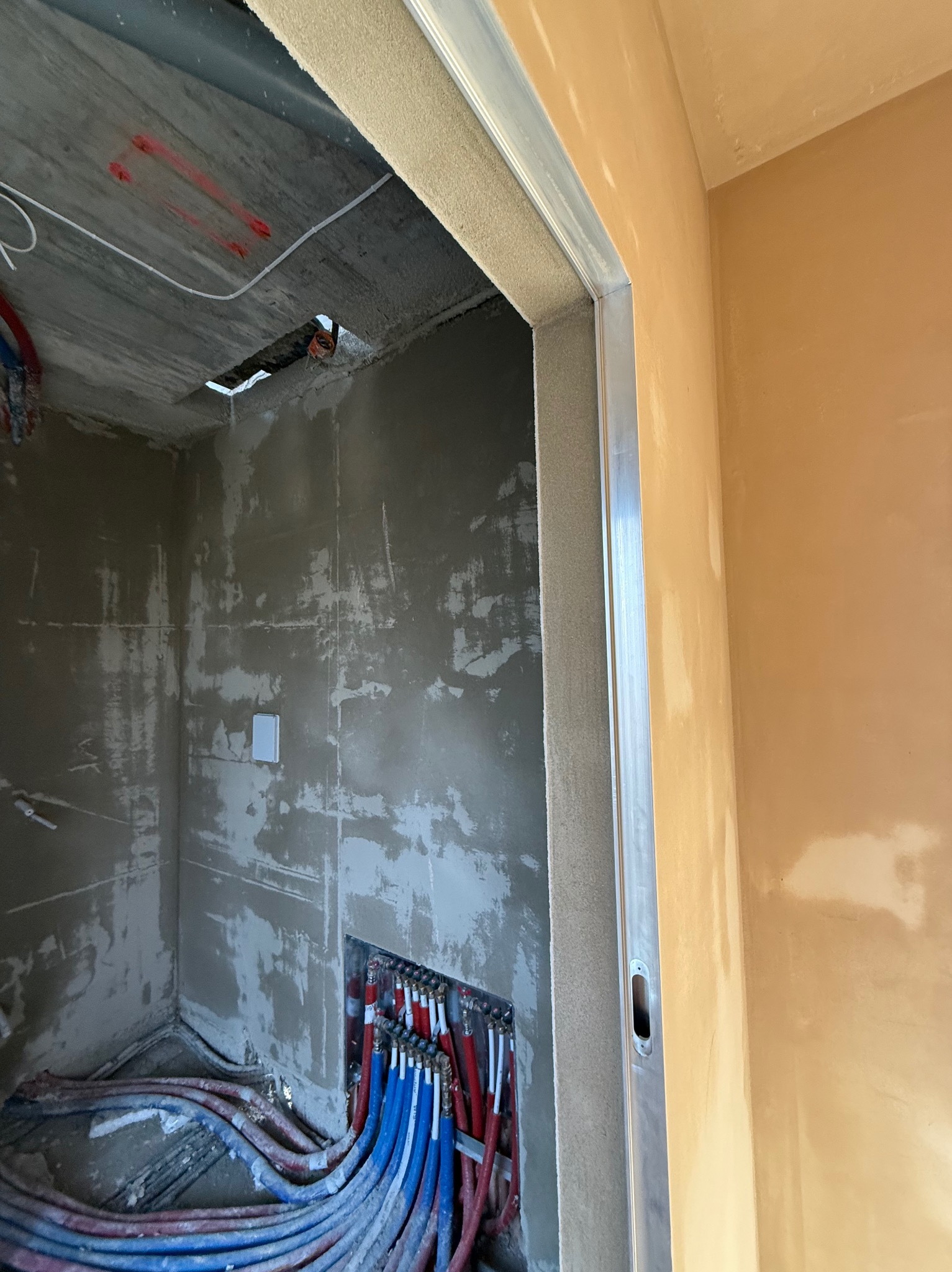 Wnętrze w trakcie prac wykończeniowych: ściany z surowym, szarym tynkiem, widoczne instalacje wodne w kolorze czerwonym i niebieskim oraz fragment ściany pokrytej jasnym, beżowym tynkiem...