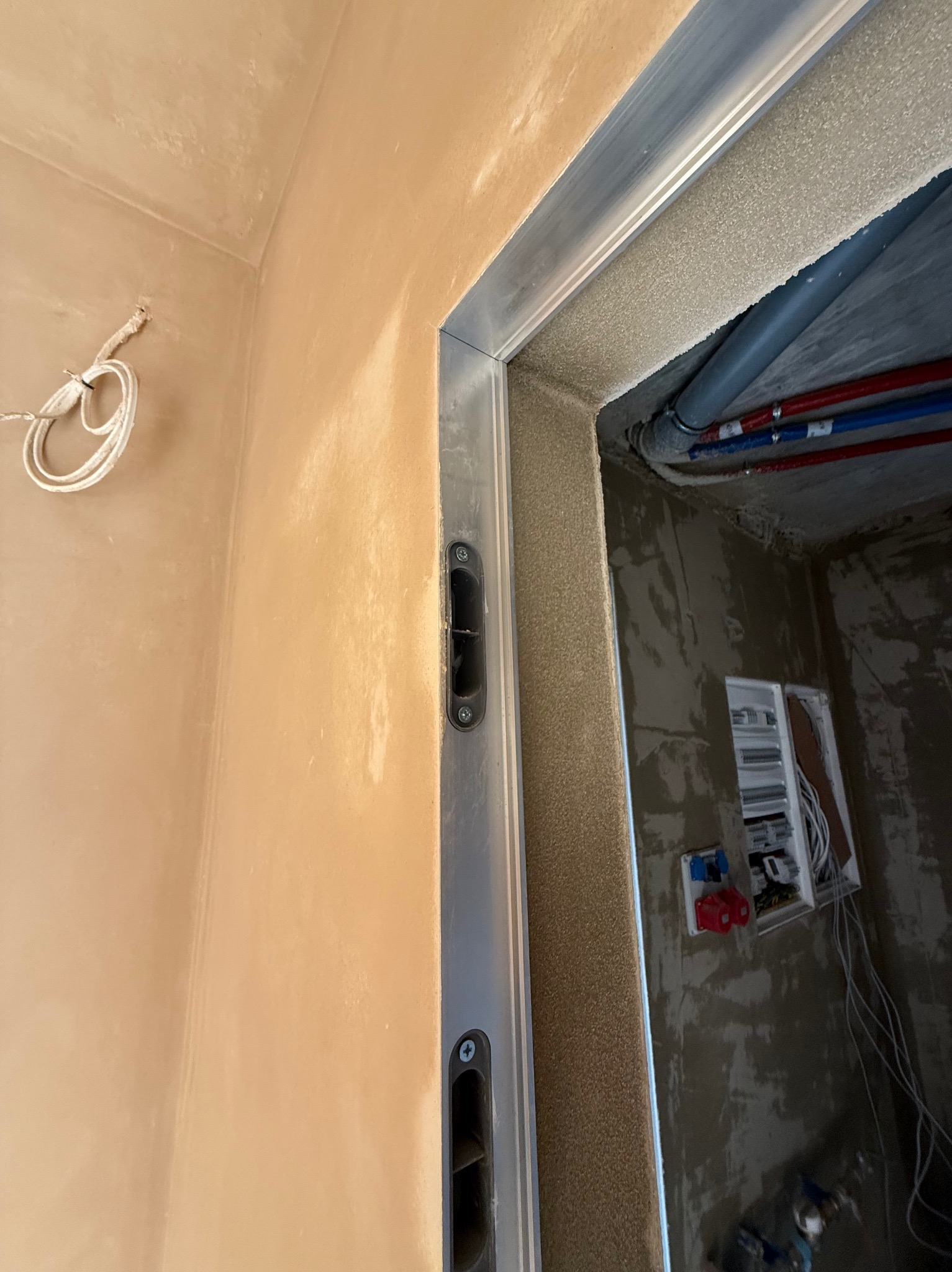 Świeżo nałożony, beżowy tynk na ścianie w korytarzu, widoczna metalowa ościeżnica z zamkiem oraz instalacje elektryczne i hydrauliczne w tle.