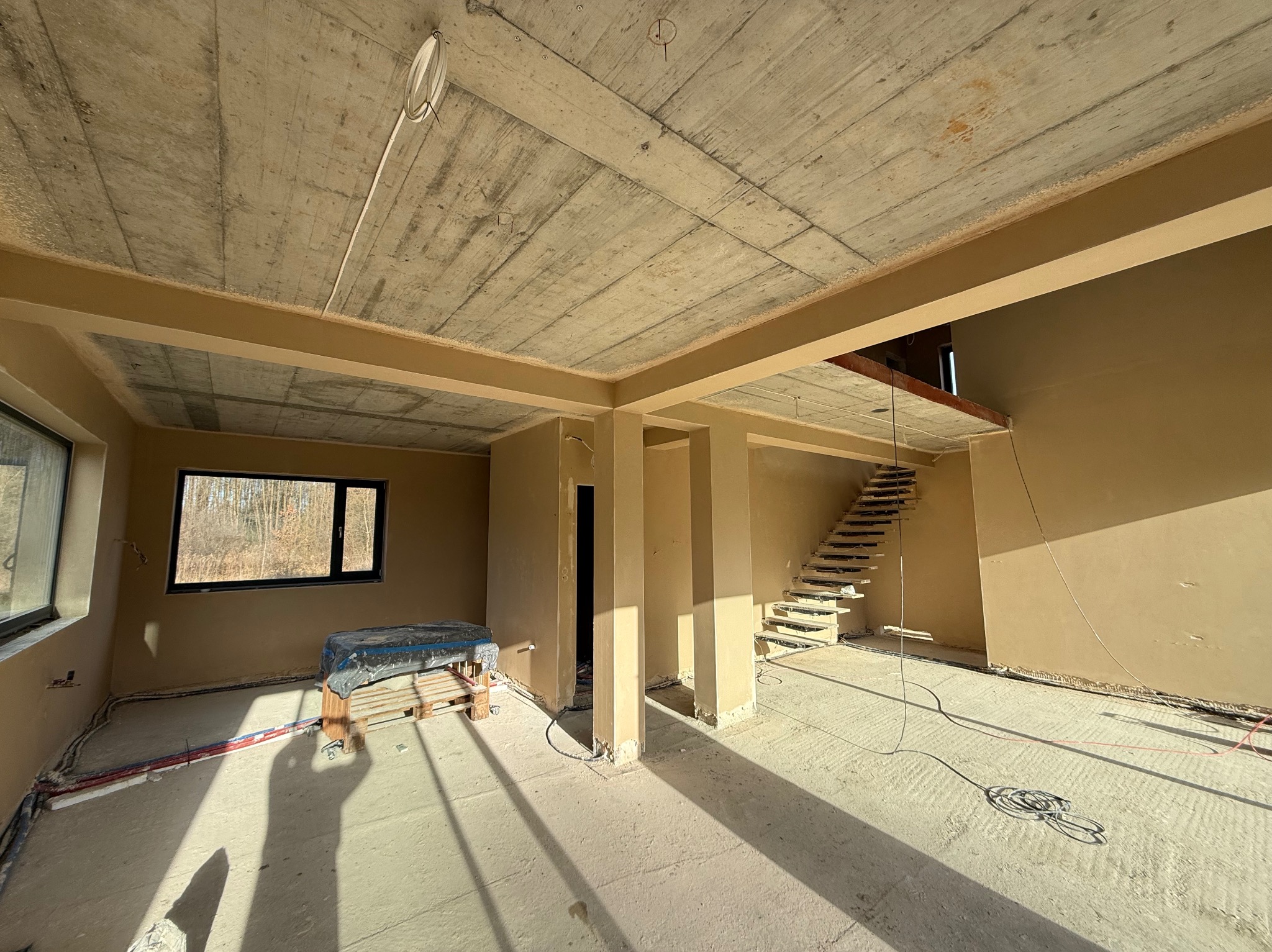 Surowy stan wnętrza domu w Krakowie po wstępnych pracach tynkarskich, widoczne ściany pokryte jasnym tynkiem, surowy betonowy strop, schody w stanie surowym oraz instalacje elektryczne...