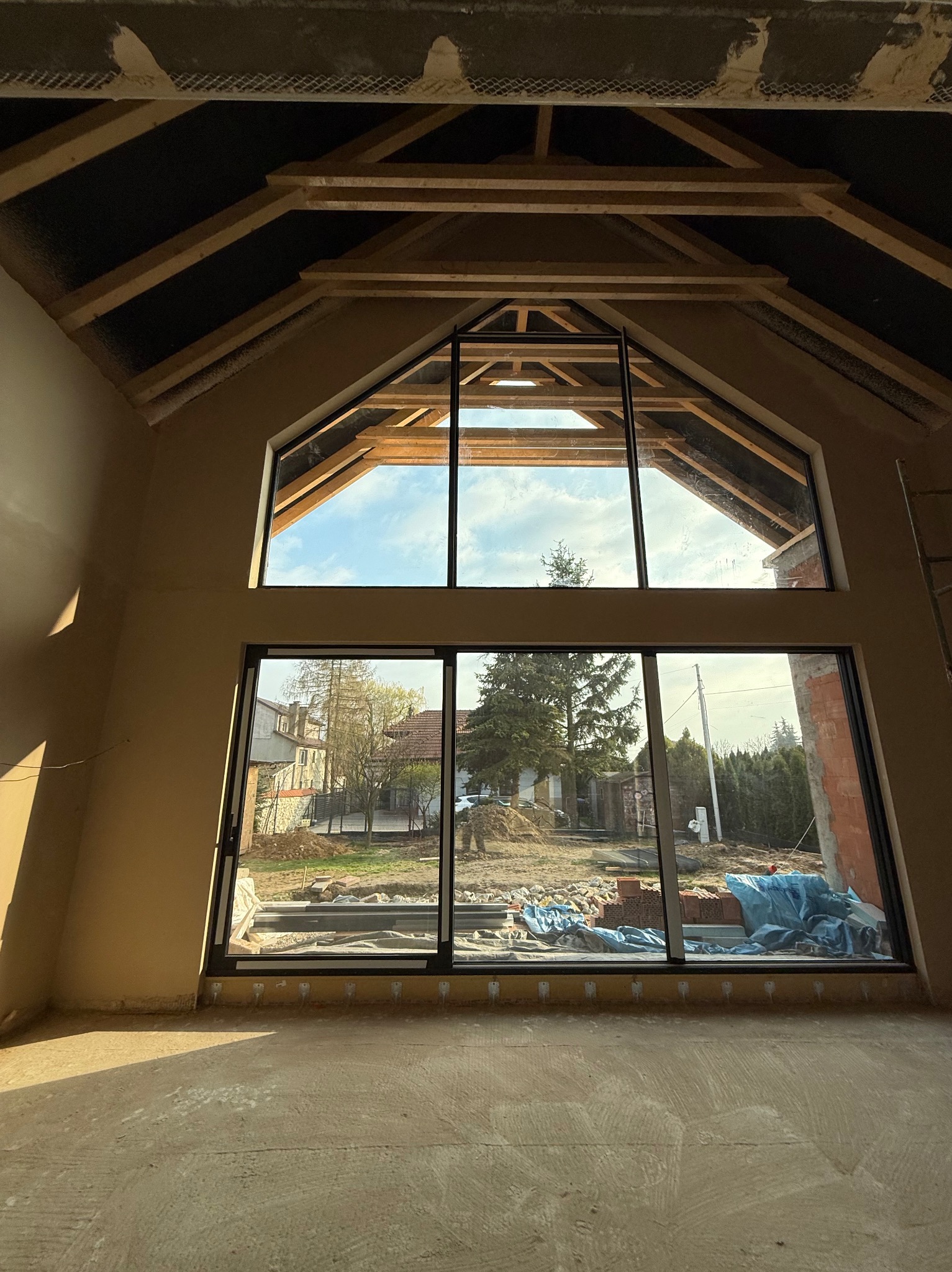 Wnętrze surowego domu z dużym oknem z widokiem na posesję, ściany pokryte świeżym tynkiem, widoczna konstrukcja dachu.