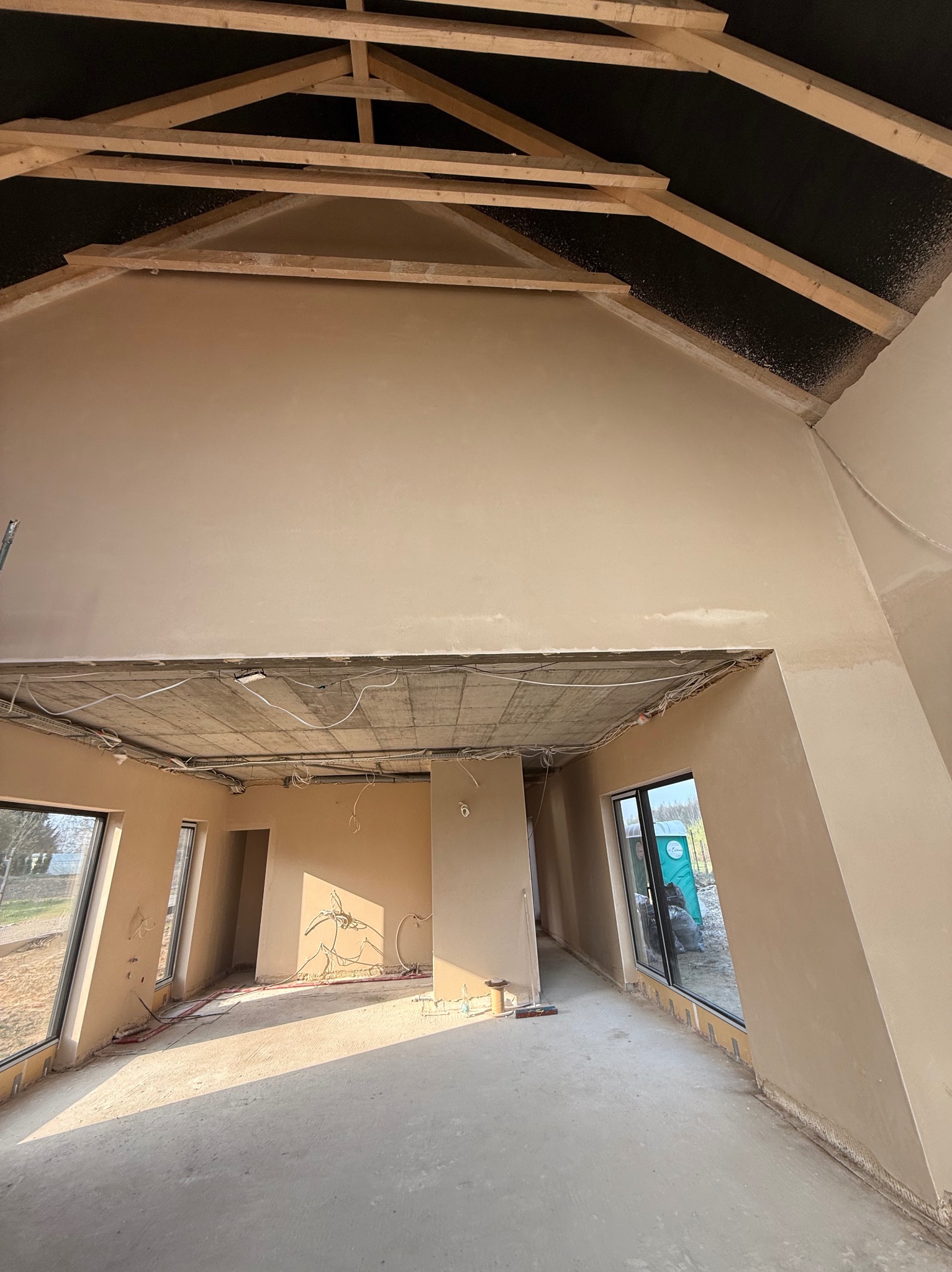 Wnętrze domu w stanie deweloperskim po nałożeniu gładzi, widoczne surowe drewniane elementy konstrukcji dachu, instalacje pod sufitem i okna z widokiem na zewnątrz.