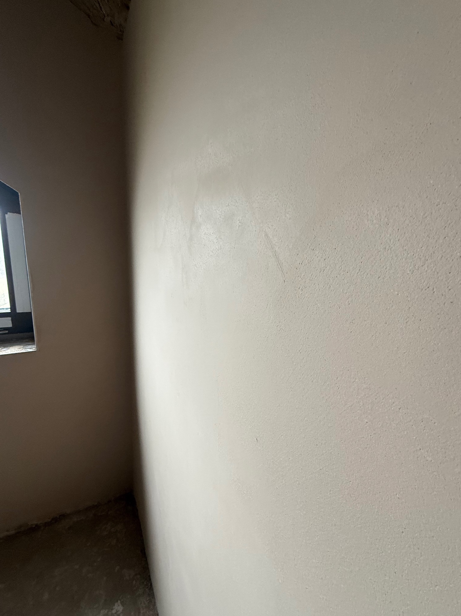 Świeżo nałożony, jasnoszary tynk maszynowy na ścianie w rogu pomieszczenia, widoczne ślady po narzędziach, okno w lewym górnym rogu kadru.