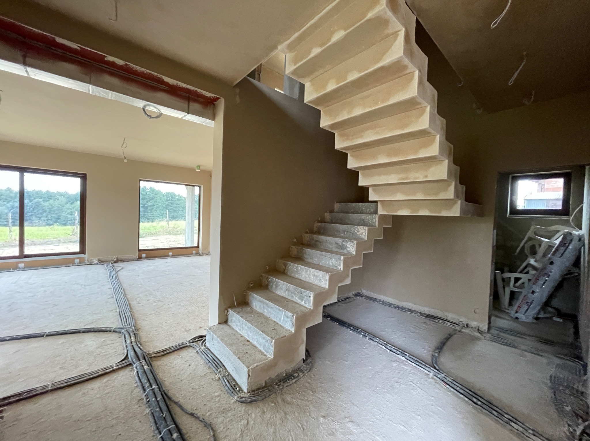 Wnętrze w stanie deweloperskim: surowe schody betonowe bez balustrady, ściany po nałożeniu gładzi, instalacje elektryczne ułożone w podłodze, widok na okno z zielenią w tle.