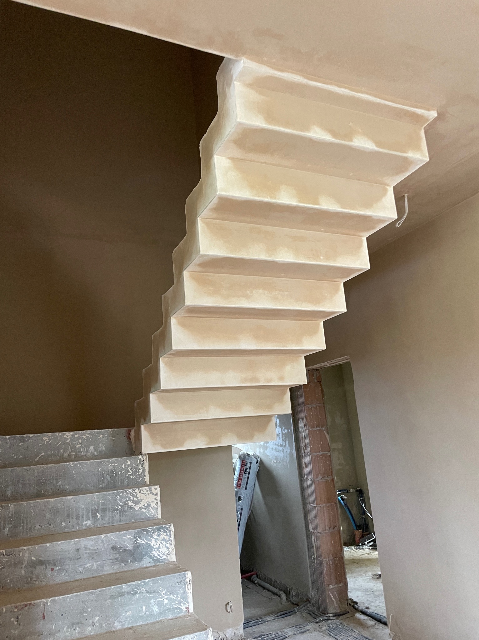 Surowe schody betonowe z nałożonym tynkiem gipsowym, widoczne niedoskonałości i nierówności, w tle nieotynkowana ściana z cegły i instalacje hydrauliczne.