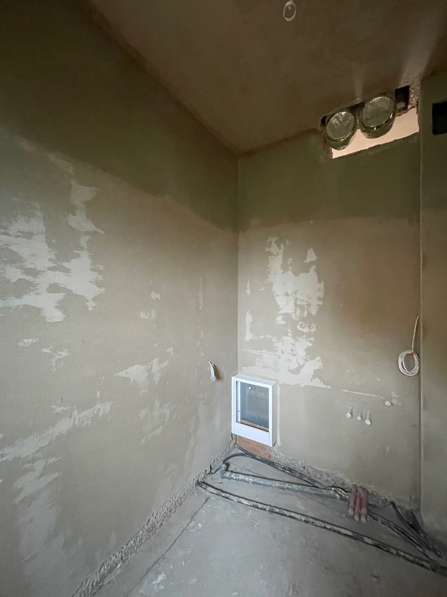 Świeżo nałożone tynki maszynowe na ścianach w surowym wnętrzu, widoczne kable elektryczne przy podłodze i otwór z lampami halogenowymi w suficie.