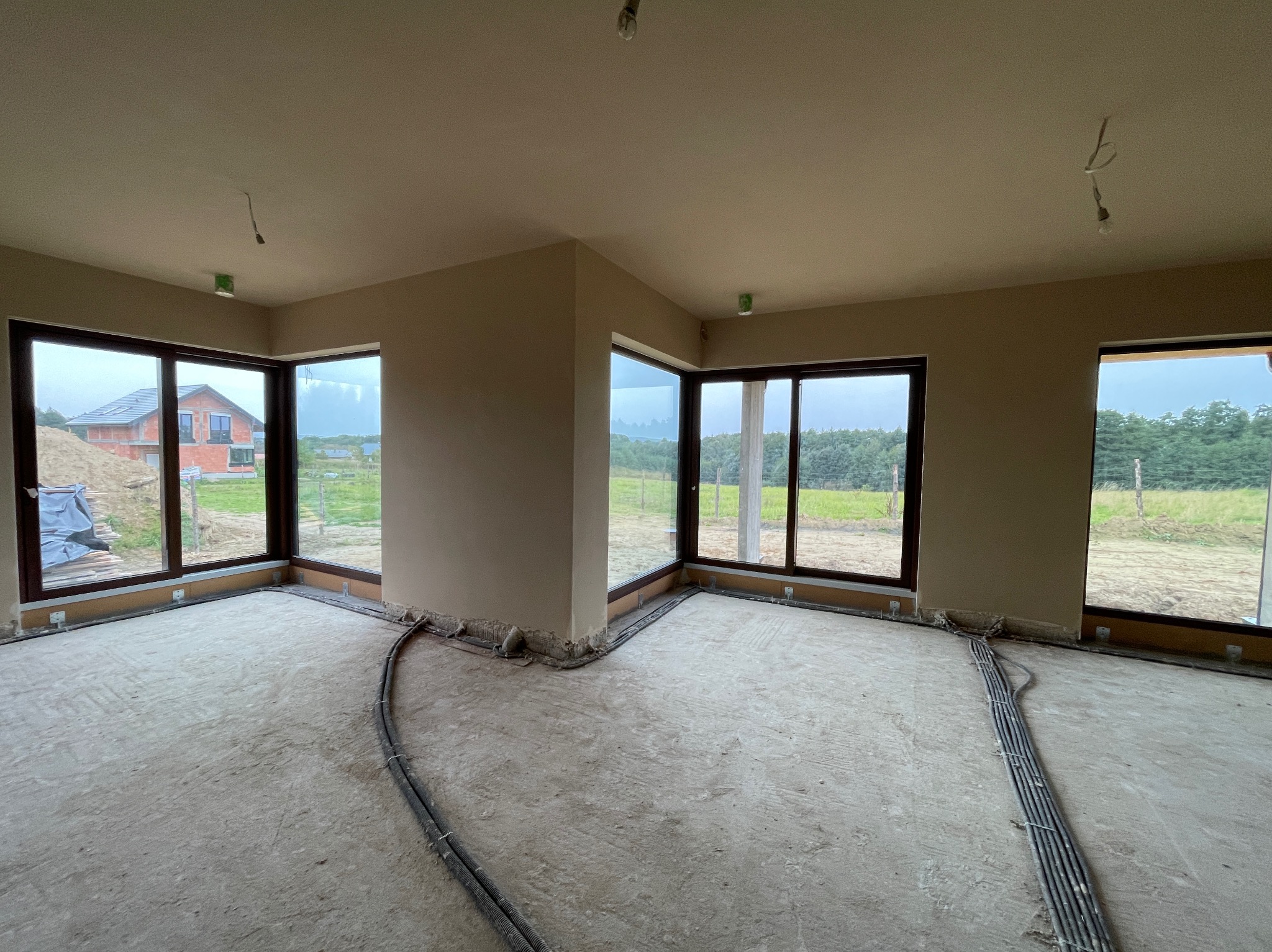 Wnętrze pomieszczenia po nałożeniu gładzi gipsowej na ścianach, widoczne okna z brązowymi ramami, wystające kable elektryczne przy podłodze i surowa podłoga.