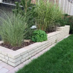 Podwyższona rabata z jasnych cegieł, obsadzona ozdobnymi trawami i niskimi krzewami, obok trawnik. Element małej architektury ogrodowej.