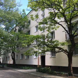 Fasada budynku wielorodzinnego z beżową elewacją, widoczna z poziomu chodnika, zieleń drzew zasłania częściowo widok, charakterystyczne okna z białymi ramami.