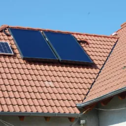 Czerwony dach z dachówki ceramicznej z zamontowanymi dwoma płaskimi panelami słonecznymi i małym panelem fotowoltaicznym. Widoczne okno dachowe i orynnowanie.