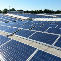 Rozległa instalacja paneli fotowoltaicznych na płaskim dachu budynku, widok z góry, błękitne niebo w tle. Panele w równych rzędach, z połyskiem odbijającym światło.