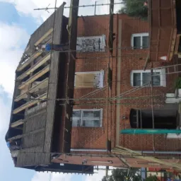 Pracownicy budowlani na rusztowaniu podczas prac przy drewnianej konstrukcji dachu na ceglanym budynku, widoczne niebo i fragment samochodu ciężarowego