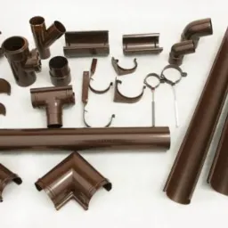 Komplet brązowych elementów systemu odprowadzania wody deszczowej: rynny, kolanka, złączki i uchwyty, rozłożone na jasnym tle.